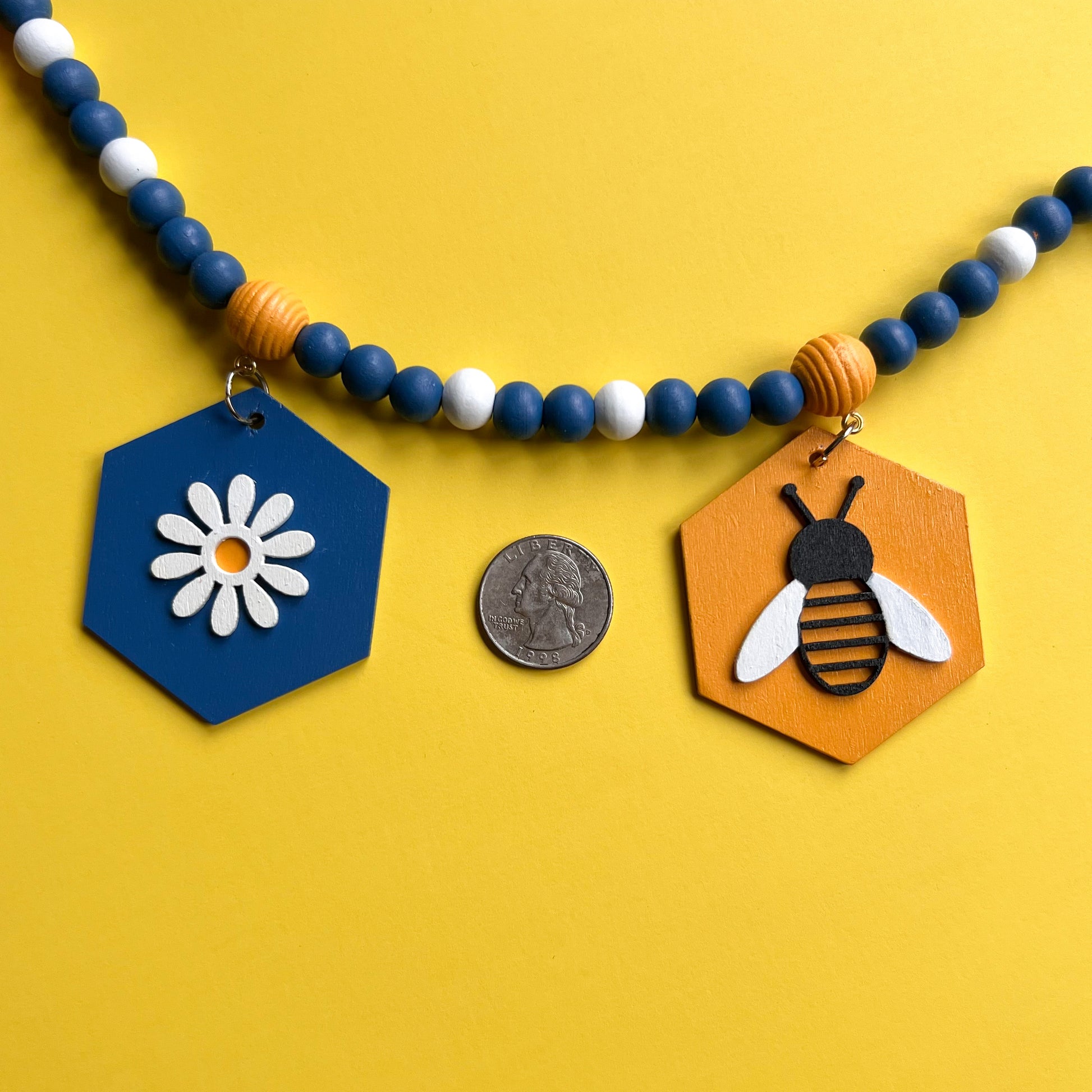 The image shows a closeup of a wood bead garland featuring wood hexagons with bees and flowers, strung with beads painted deep blue, golden yellow, and white. Hexagons hang from beehive shaped beads. Beads are strung on 2mm blue macrame cord with 3-inch loops on each end.