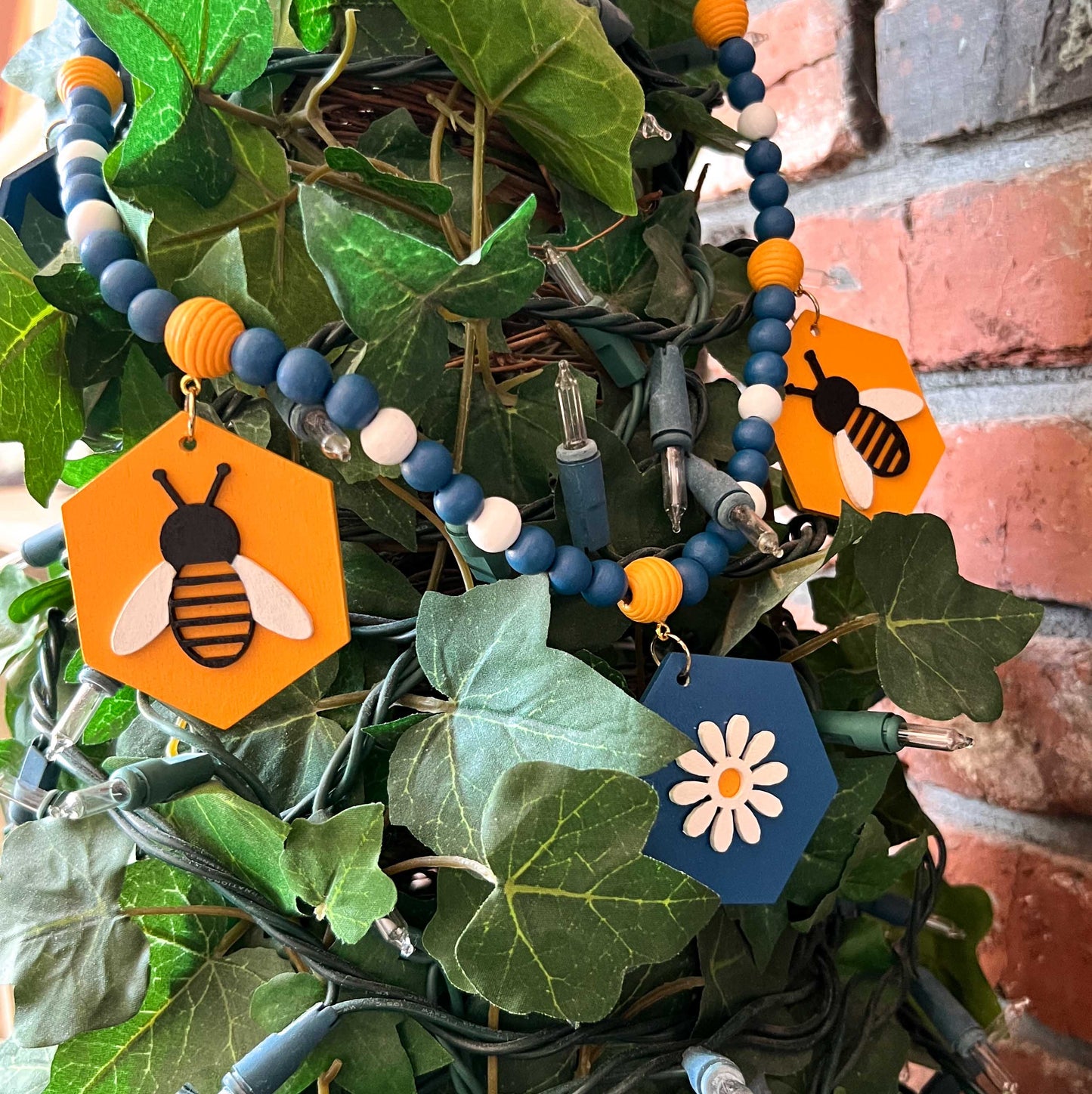 The image shows a wood bead garland featuring wood hexagons with bees and flowers, strung with beads painted deep blue, golden yellow, and white. Hexagons hang from beehive shaped beads. Beads are strung on 2mm blue macrame cord with 3-inch loops on each end.