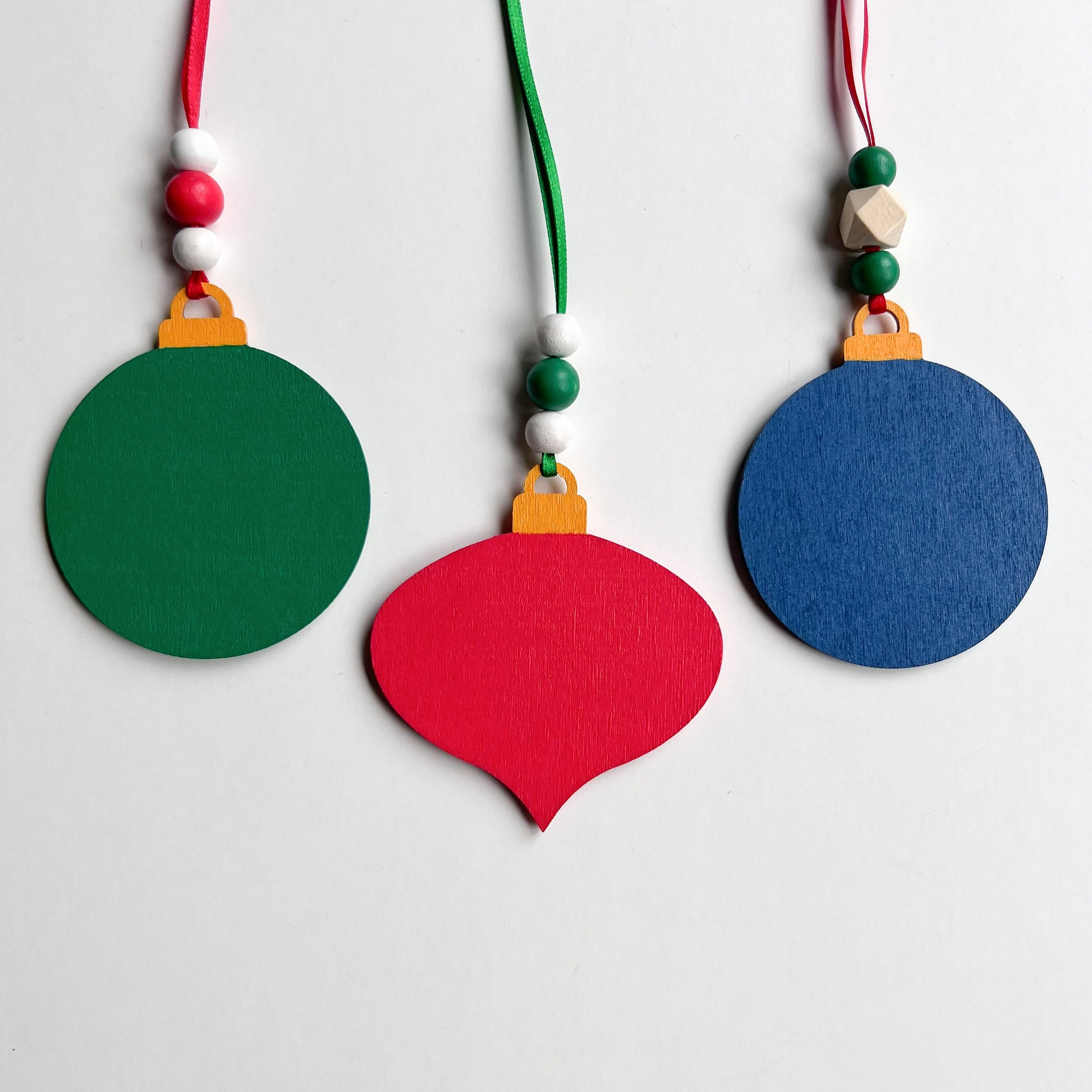 Image shows back of a set of 3 flat wood handmade ornaments. The front of each ornament features a holiday themed wood shape: a decorated Christmas tree, a red stocking, or a truck bringing home the tree. Ornaments hang from ribbon and are decorated with three wood beads in matching colors.