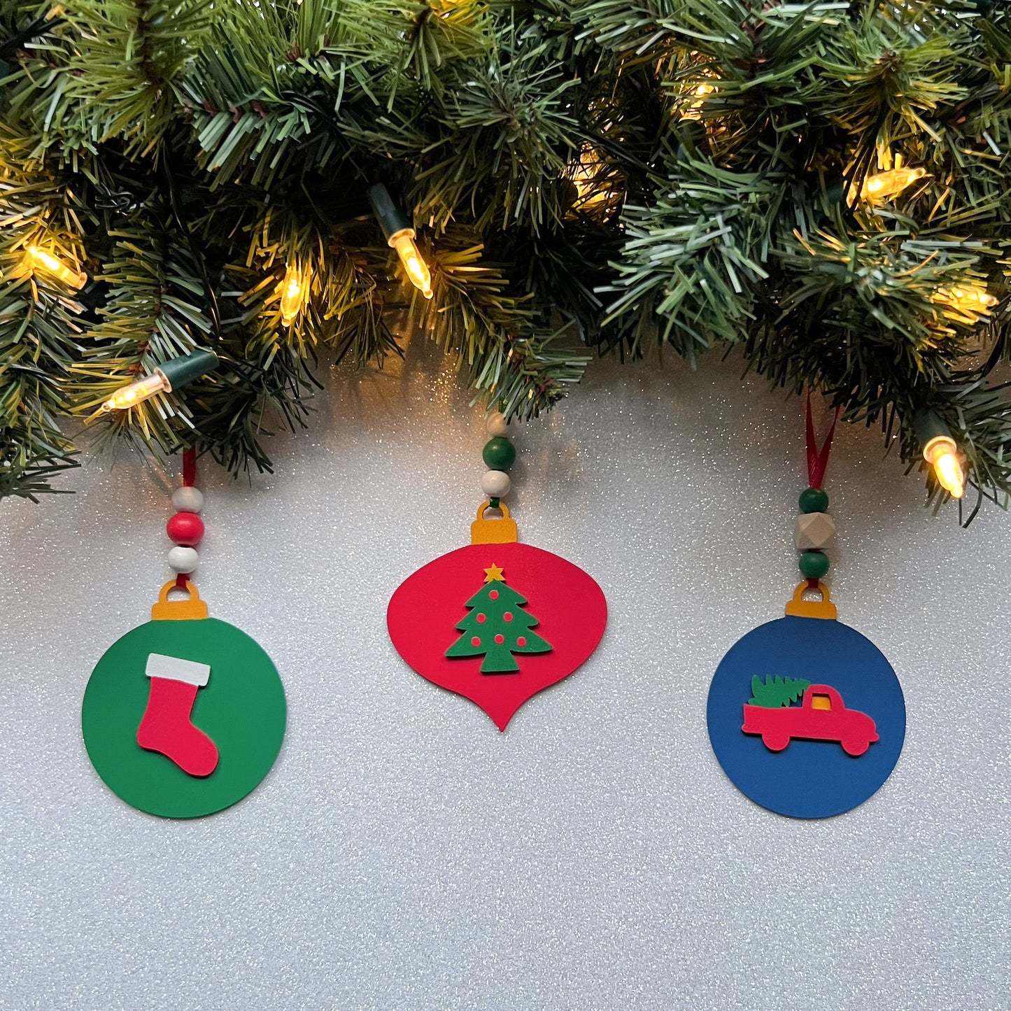 Image shows set of 3 flat wood handmade ornaments. Each ornament features a holiday themed wood shape: a decorated Christmas tree, a red stocking, or a truck bringing home the tree. Ornaments hang from ribbon and are decorated with three wood beads in matching colors.