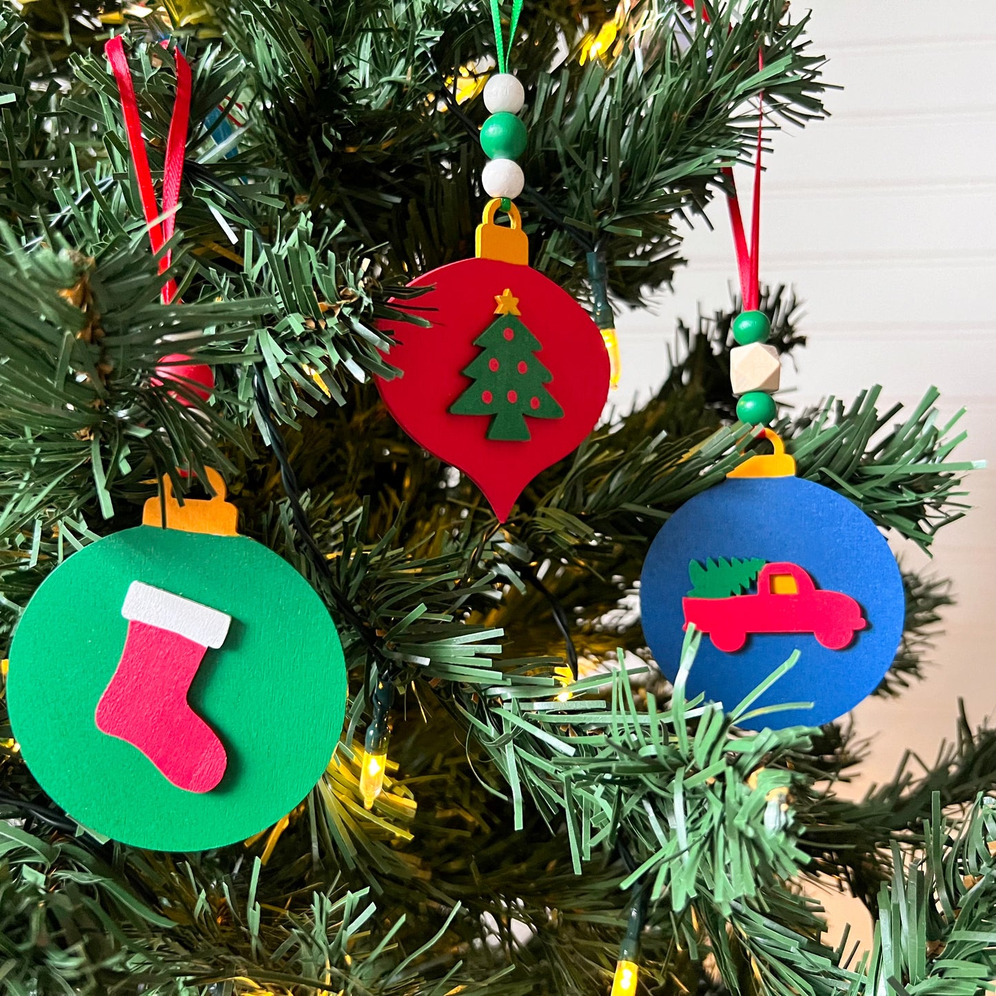 Image shows set of 3 flat wood handmade ornaments. Each ornament features a holiday themed wood shape: a decorated Christmas tree, a red stocking, or a truck bringing home the tree. Ornaments hang from ribbon and are decorated with three wood beads in matching colors.