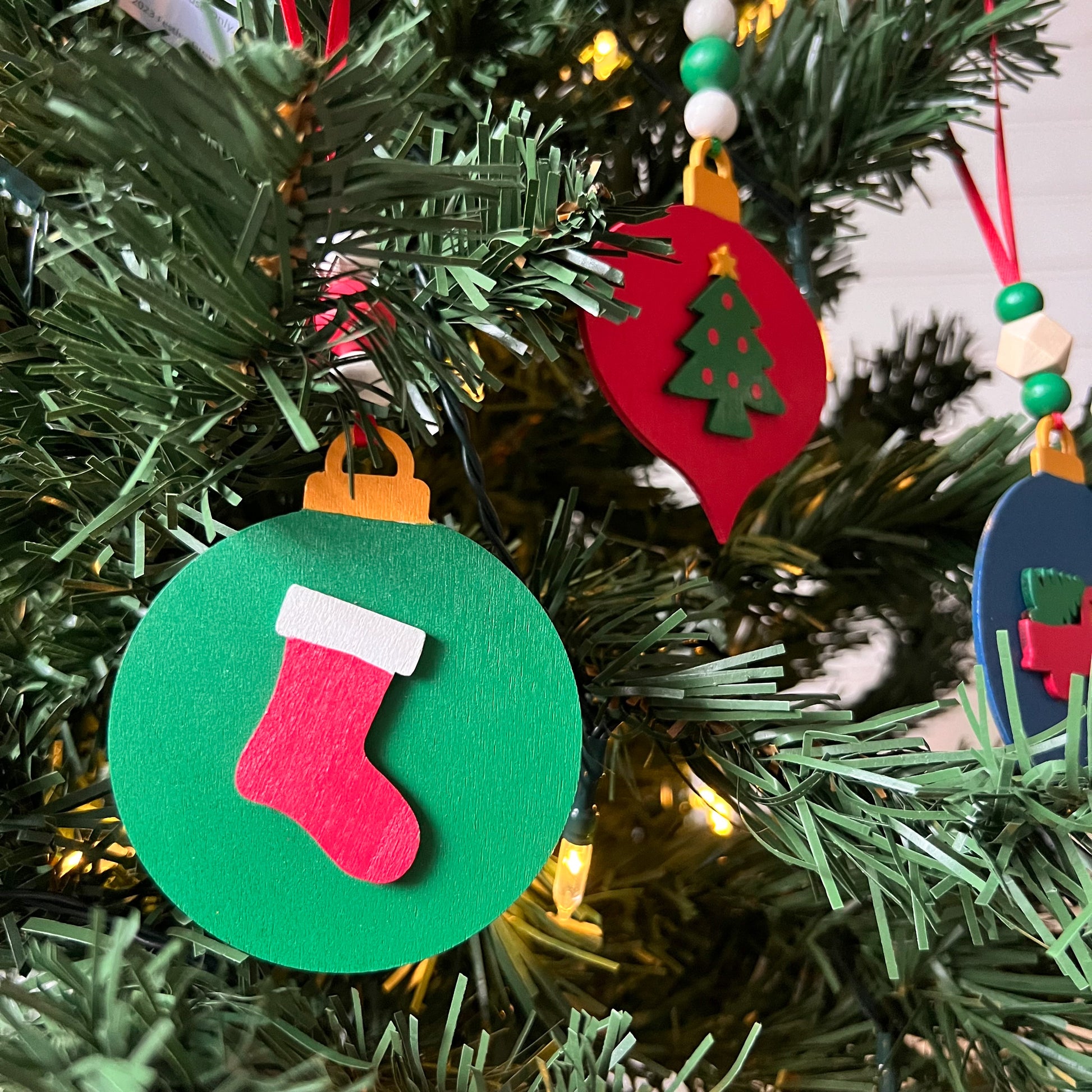 Image shows set of 3 flat wood handmade ornaments. Each ornament features a holiday themed wood shape: a decorated Christmas tree, a red stocking, or a truck bringing home the tree. Ornaments hang from ribbon and are decorated with three wood beads in matching colors.