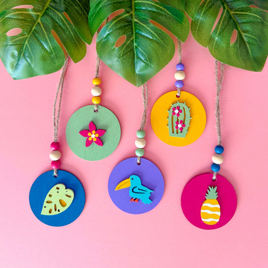 Small 2 inch round wood ornaments with tropical themed shapes including a pineapple, a cactus, a tropical leaf, a flower, and a bird. Ornaments hang from jute cording with three small beads. Painted in bright colors of magenta, yellow, lavender, green, and medium blue.