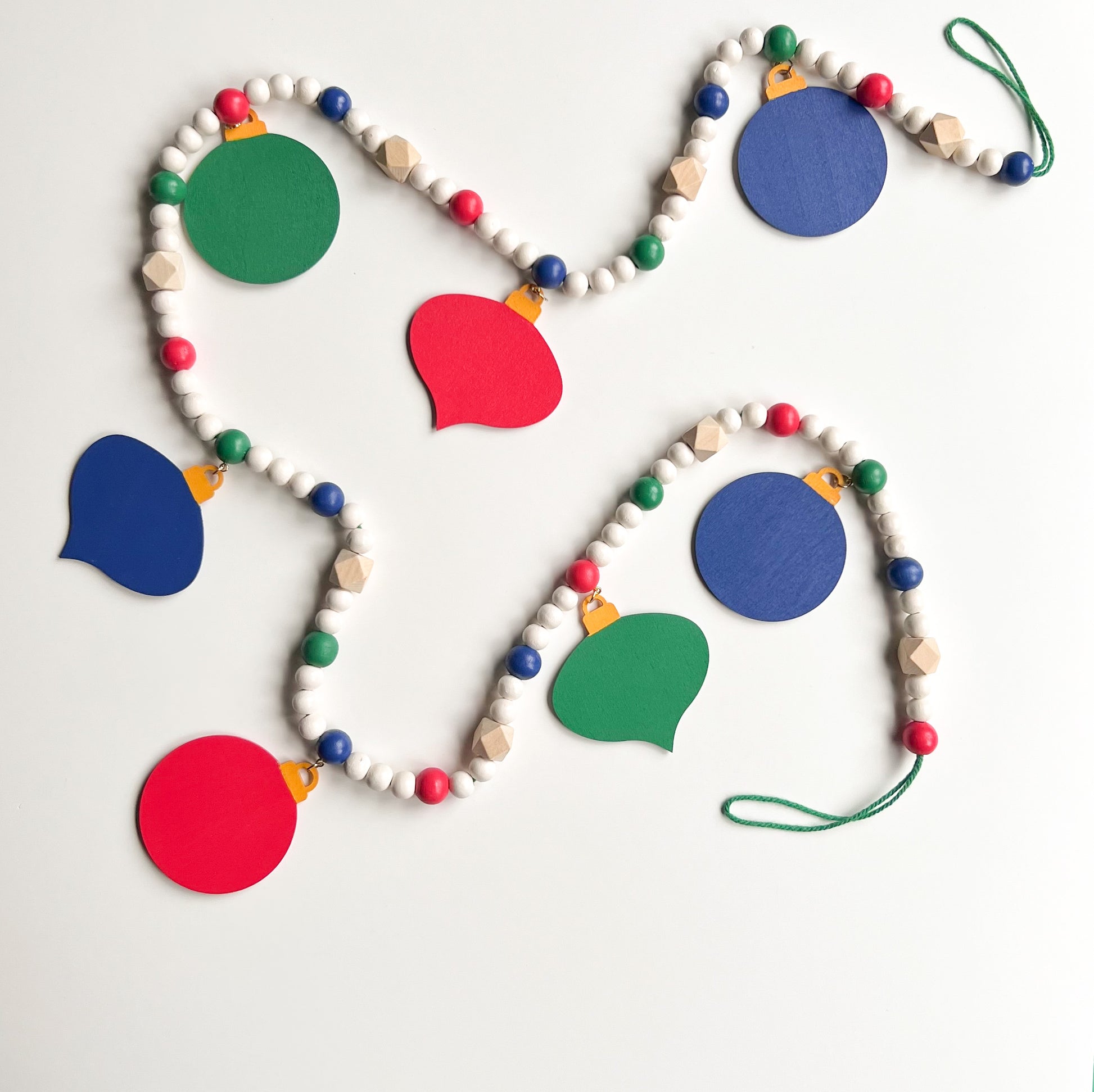 Back of Christmas themed wood bead garland with 7 flat wood ornaments hanging from wood beads. Ornaments feature different holiday shapes. Colors in beads and ornaments are green, blue, red, white, and yellow. Garland is on a white background.