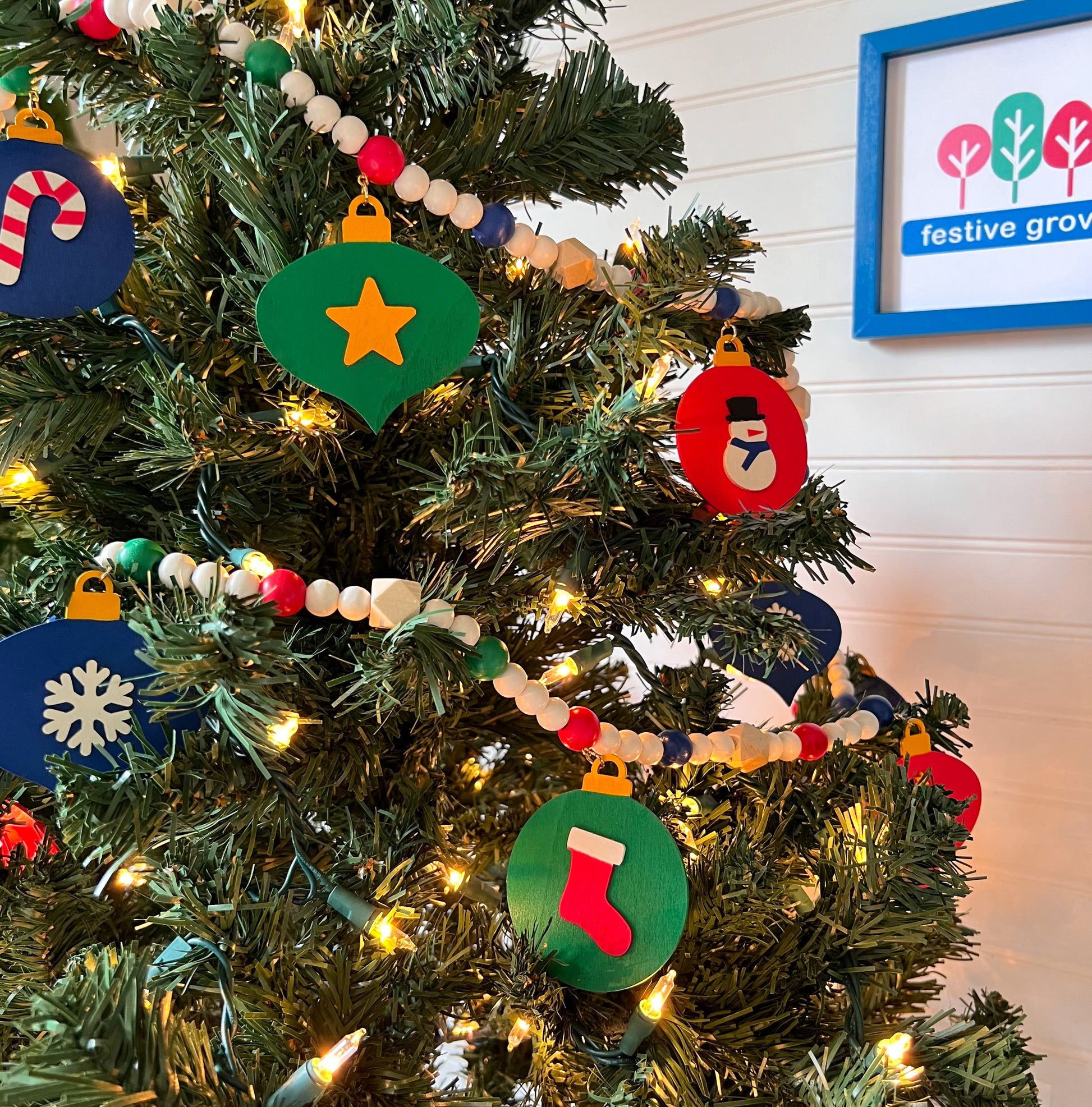 Christmas themed wood bead garland with 7 flat wood ornaments hanging from wood beads. Ornaments feature different holiday shapes. Colors in beads and ornaments are green, blue, red, white, and yellow. Garland is on a Christmas tree..