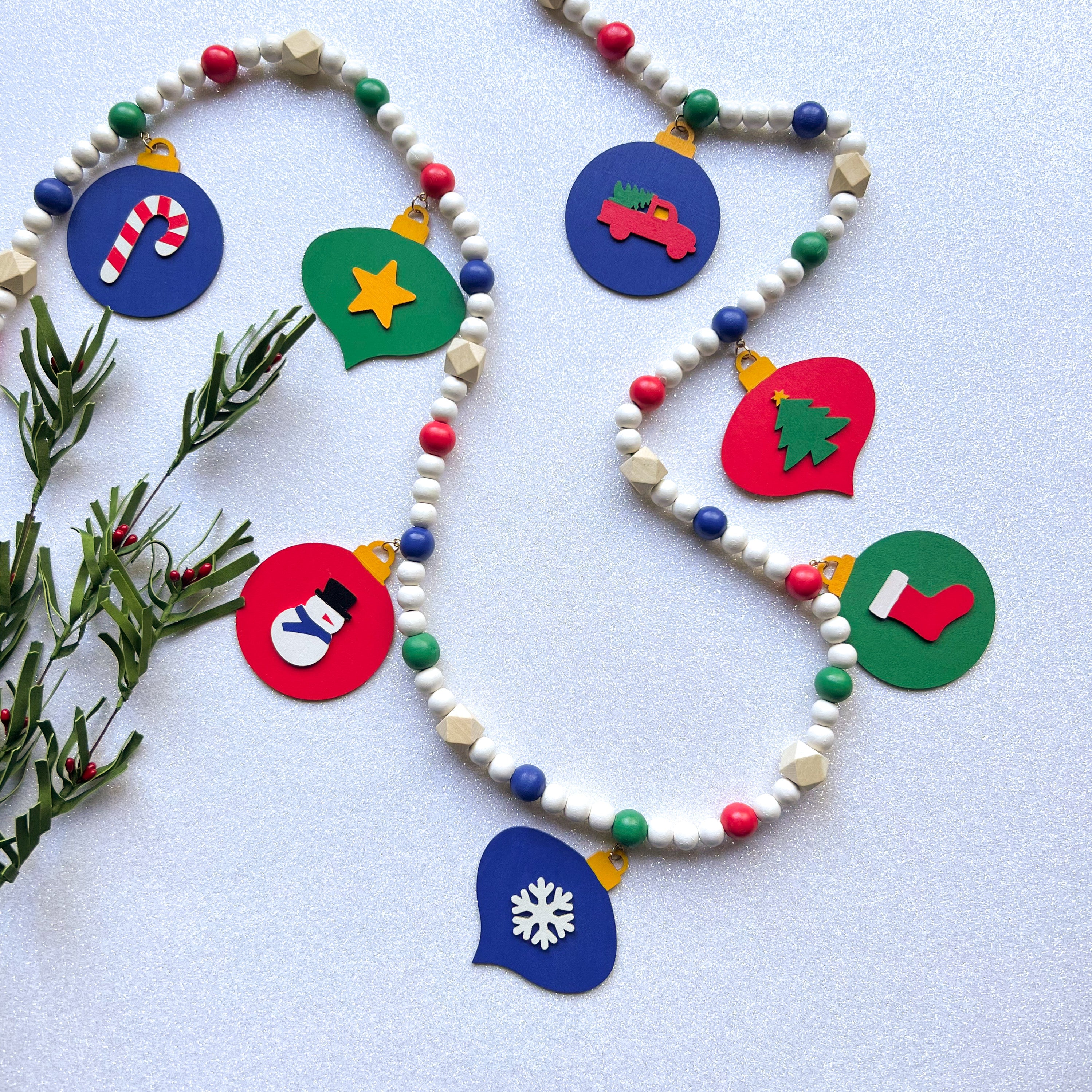 Christmas themed wood bead garland with 7 flat wood ornaments hanging from wood beads. Ornaments feature different holiday shapes. Colors in beads and ornaments are green, blue, red, white, and yellow. Garland is on a white glitter background.