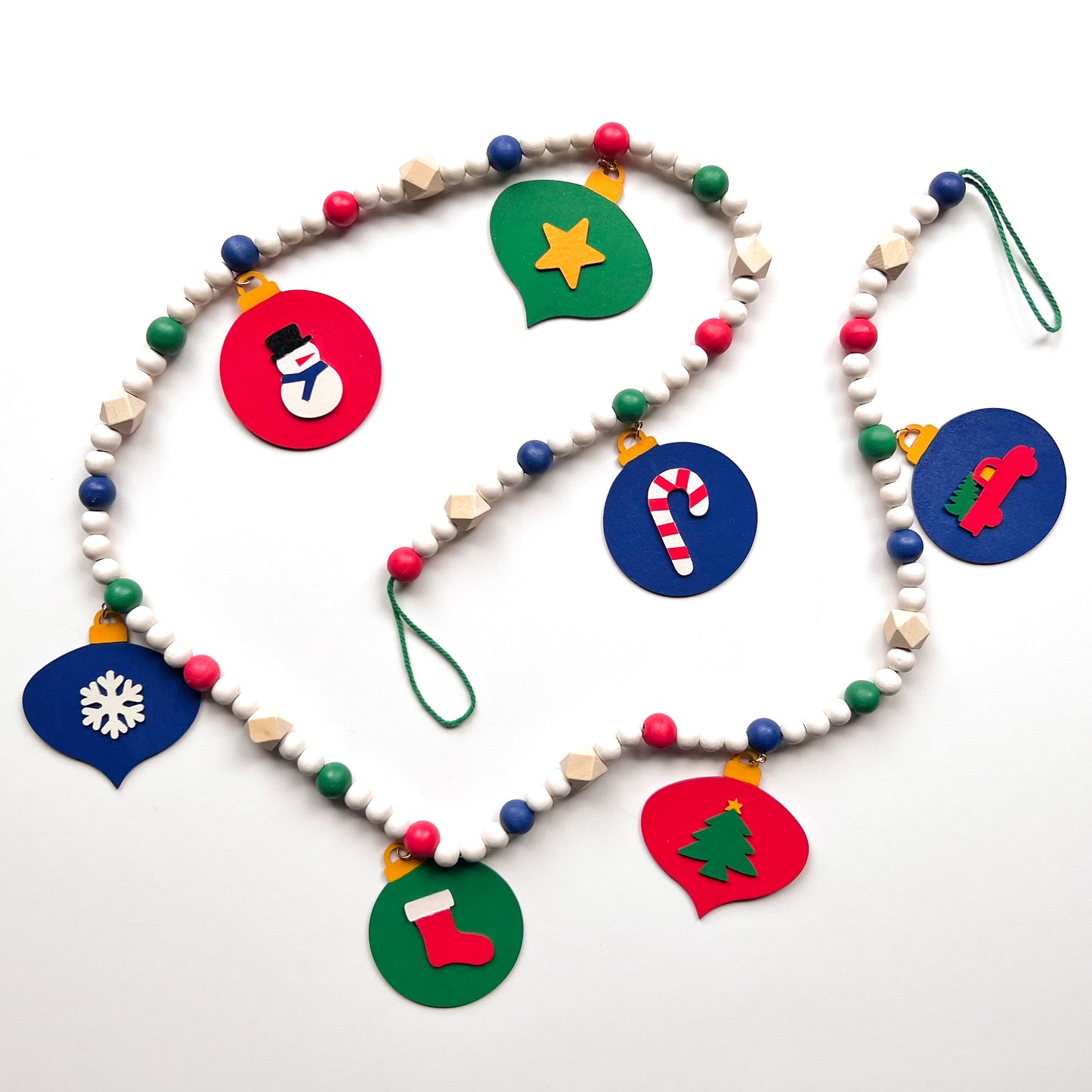 Christmas themed wood bead garland with 7 flat wood ornaments hanging from wood beads. Ornaments feature different holiday shapes. Colors in beads and ornaments are green, blue, red, white, and yellow. Garland is on a white background.