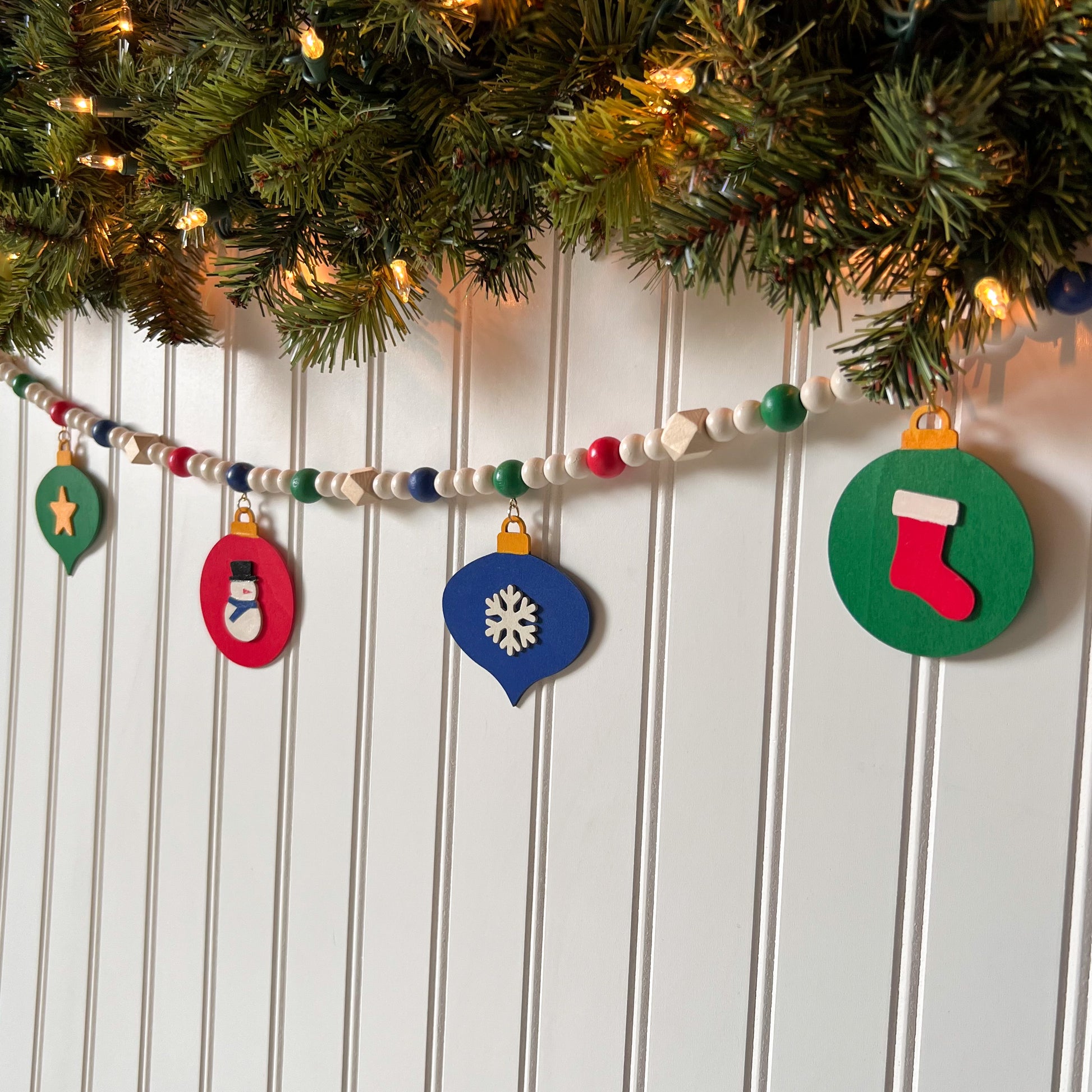 Christmas themed wood bead garland with 7 flat wood ornaments hanging from wood beads. Ornaments feature different holiday shapes. Colors in beads and ornaments are green, blue, red, white, and yellow. Garland is on a white background.