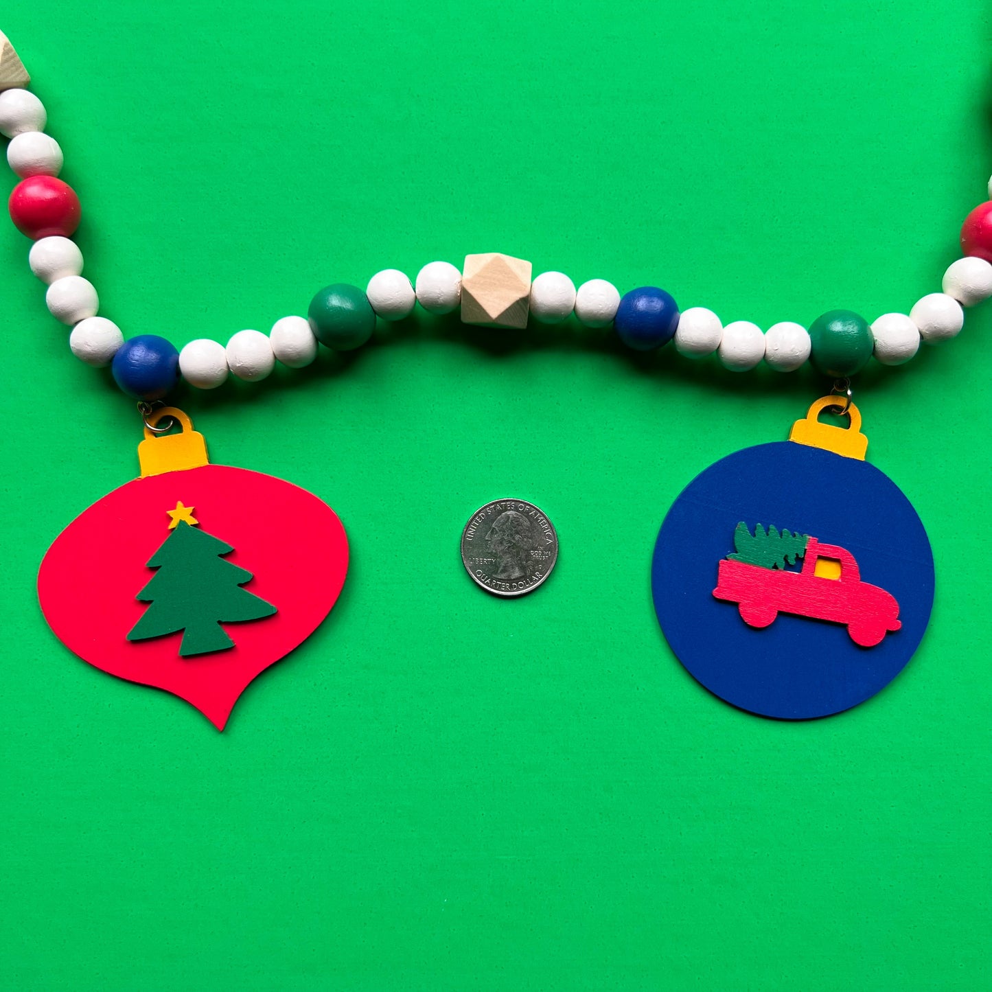 Christmas themed wood bead garland with 7 flat wood ornaments hanging from wood beads. Ornaments feature different holiday shapes. Colors in beads and ornaments are green, blue, red, white, and yellow. Garland is on a green background.