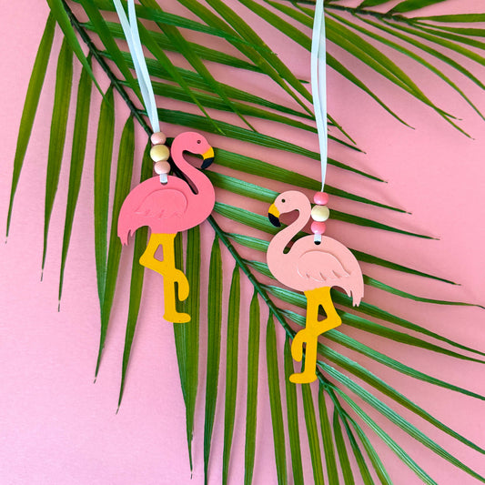 Two wood flamingo ornaments in different shades of pink hang from a 6" white ribbon with three beads in white and pink. Ornaments are on a palm leaf.