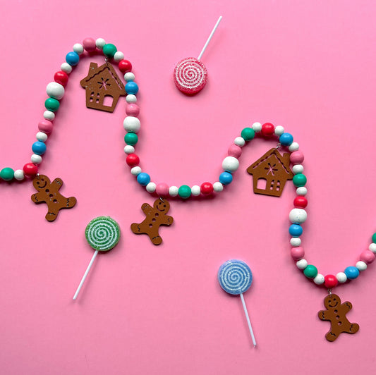 Gingerbread Village Handmade Garland. The garland features three wood gingerbread figures alongside two gingerbread houses. Shapes hang from wood beads. Christmas holiday seasonal decoration or gift.