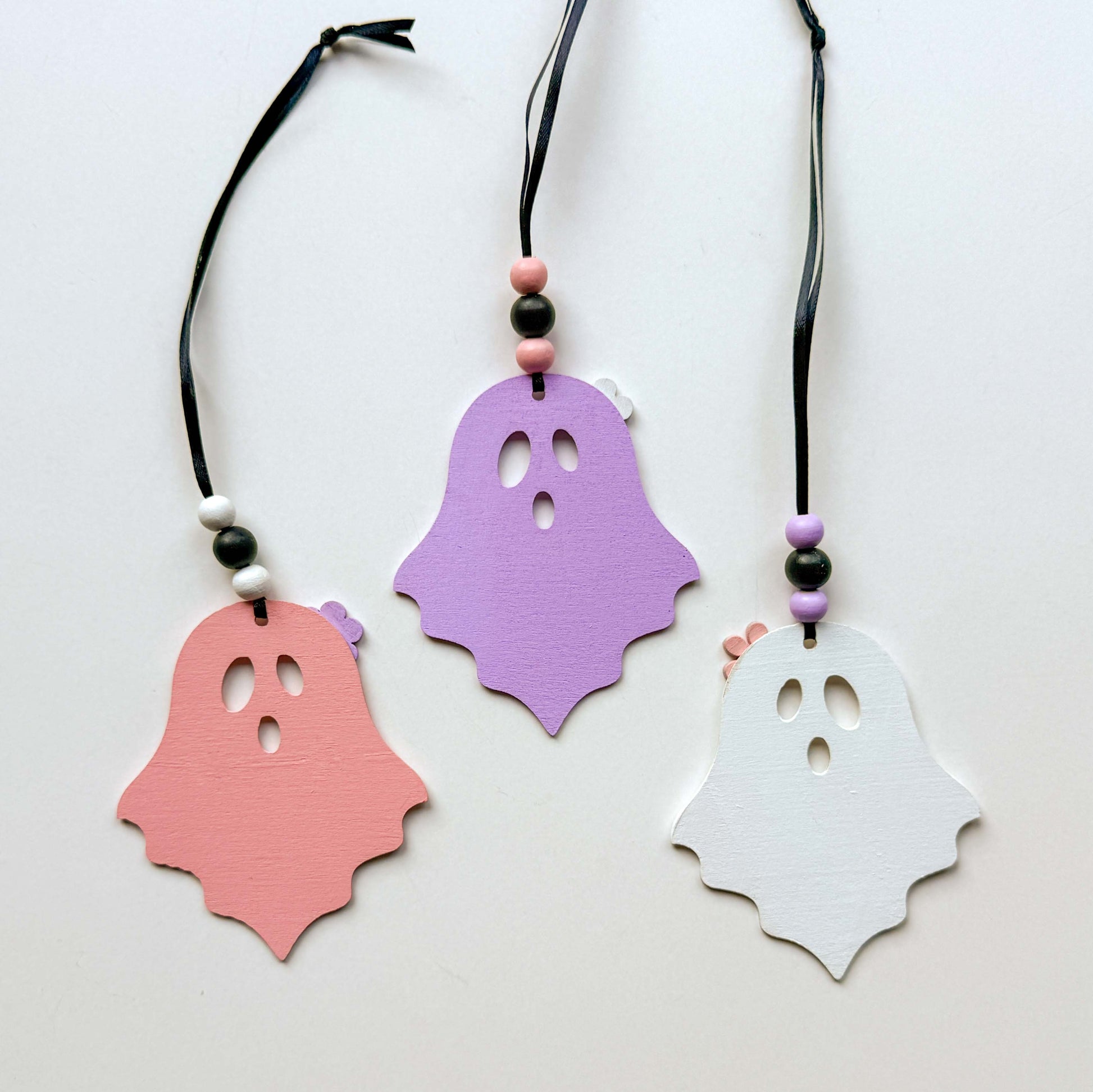 Set of three wood ghost Halloween ornaments. Three wood ghosts, in light pink, purple, or white, feature a flower accent. Ghosts hang from black ribbon; three wood beads in corresponding colors complete the ornament.