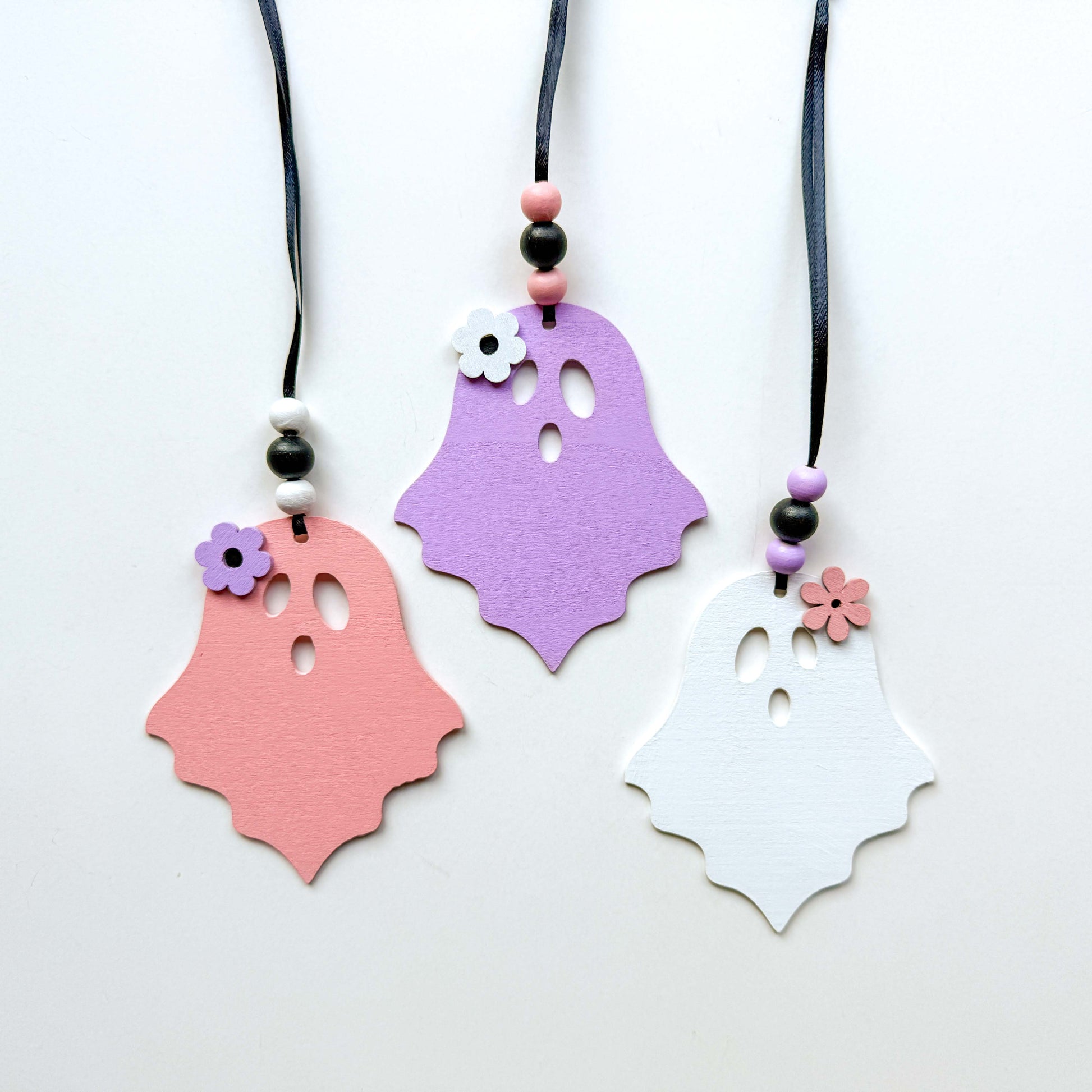 Set of three wood ghost Halloween ornaments. Three wood ghosts, in light pink, purple, or white, feature a flower accent. Ghosts hang from black ribbon; three wood beads in corresponding colors complete the ornament.