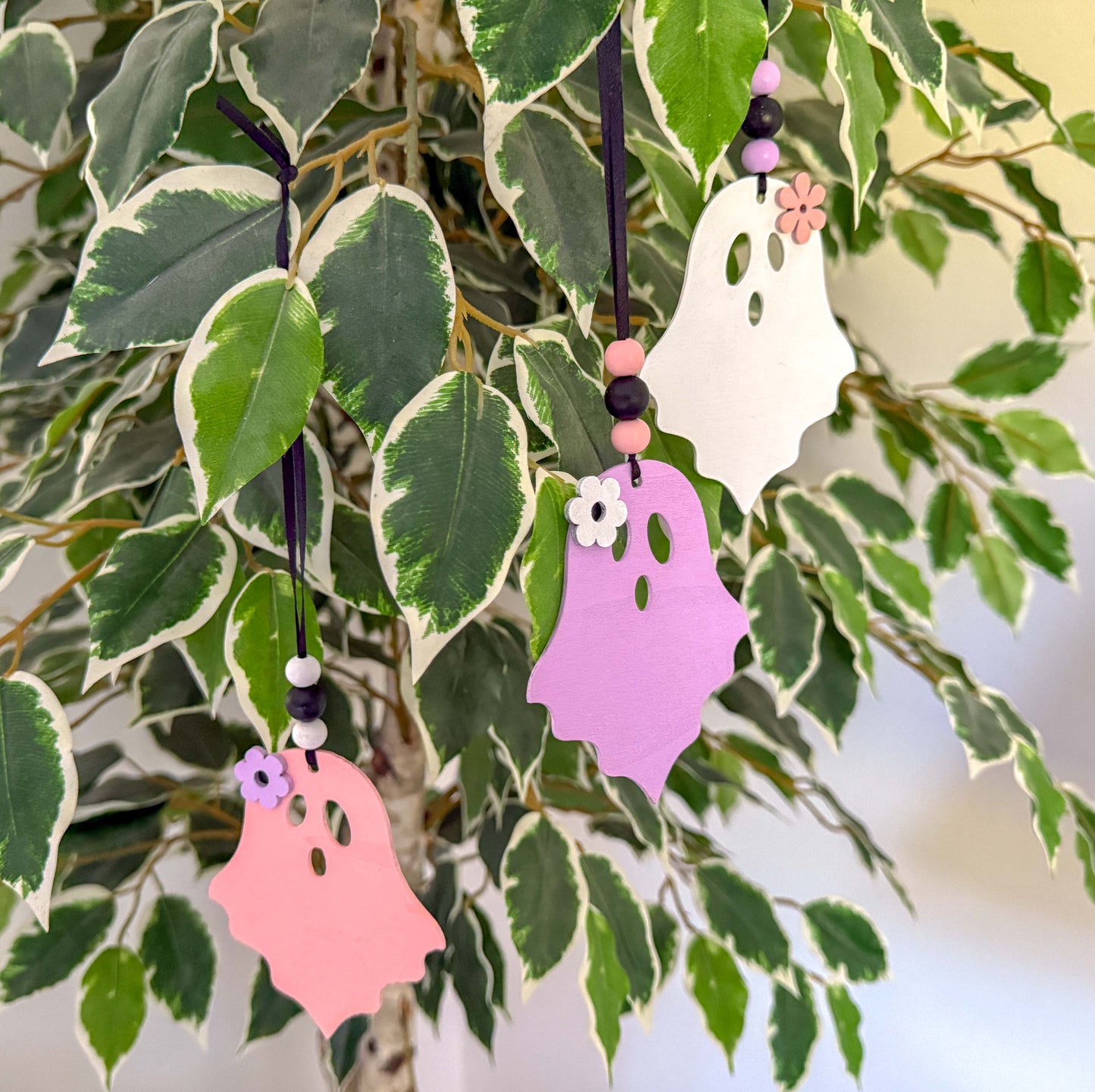 Set of three wood ghost Halloween ornaments. Three wood ghosts, in light pink, purple, or white, feature a flower accent. Ghosts hang from black ribbon; three wood beads in corresponding colors complete the ornament.