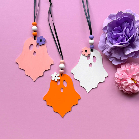 Set of three wood ghost Halloween ornaments. Three wood ghosts, in light pink, bright orange, or white, feature a flower accent. Ghosts hang from black ribbon; three wood beads in corresponding colors complete the ornament.