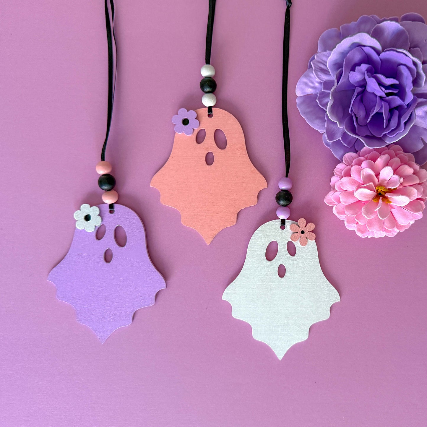 Set of three wood ghost Halloween ornaments. Three wood ghosts, in light pink, purple, or white, feature a flower accent. Ghosts hang from black ribbon; three wood beads in corresponding colors complete the ornament.