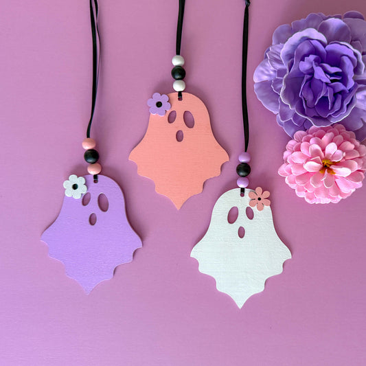 Set of three wood ghost Halloween ornaments. Three wood ghosts, in light pink, purple, or white, feature a flower accent. Ghosts hang from black ribbon; three wood beads in corresponding colors complete the ornament.