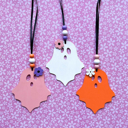 Set of three wood ghost Halloween ornaments. Three wood ghosts, in light pink, bright orange, or white, feature a flower accent. Ghosts hang from black ribbon; three wood beads in corresponding colors complete the ornament.