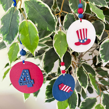 Set of 3 patriotic themed ornaments painted in colors of red, white, and blue. Ornaments feature Uncle Sam's hat,  "4th of July" cutout, and a firecracker. Ornaments hang from a ribbon with three painted wood beads.