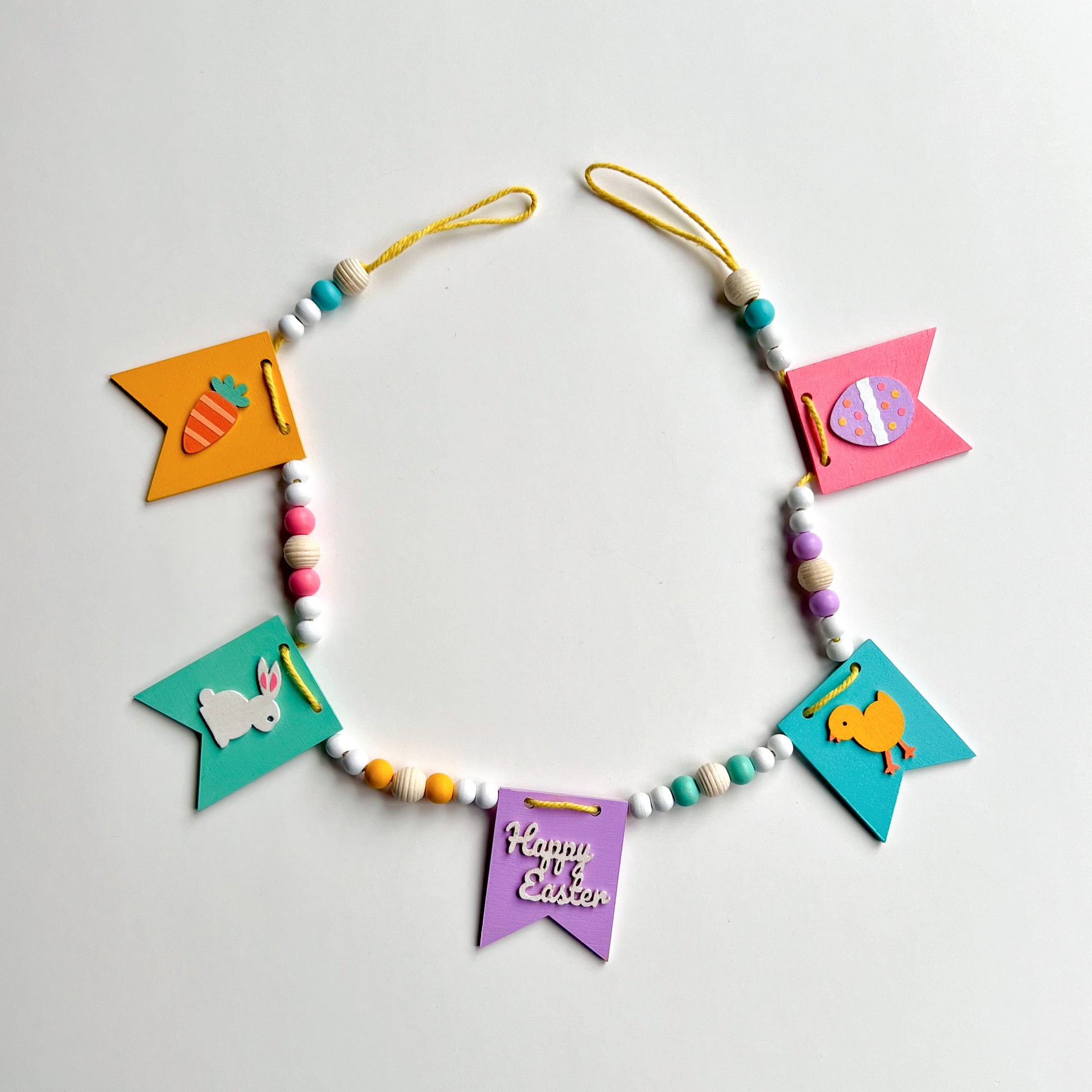 Image shows handmade garland with wood pennants featuring Easter shapes - a carrot, a bunny, a chick, an egg, and a "Happy Easter" wood cutout. Pennants are strung with natural and painted beads; includes 3-inch loops on either end. 