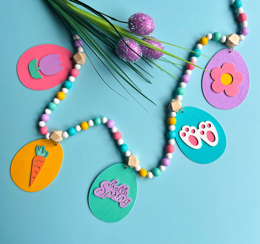 Image shows handmade garland with flat wood eggs featuring spring themed shapes and hang from a geometric wood bead. Garland has 4-inch loop on either end.