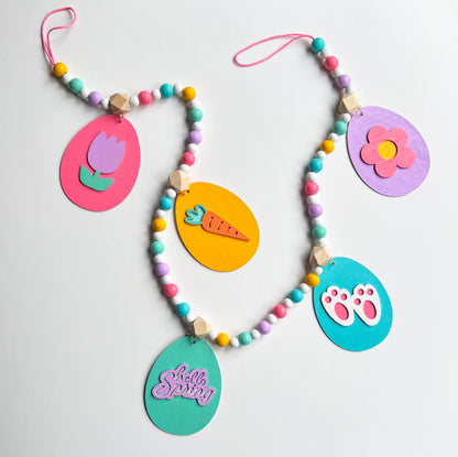 Image shows handmade garland with flat wood eggs featuring spring themed shapes and hang from a geometric wood bead. Garland has 4-inch loop on either end.