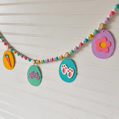 Image shows handmade garland with flat wood eggs featuring spring themed shapes and hang from a geometric wood bead. Garland has 4-inch loop on either end.