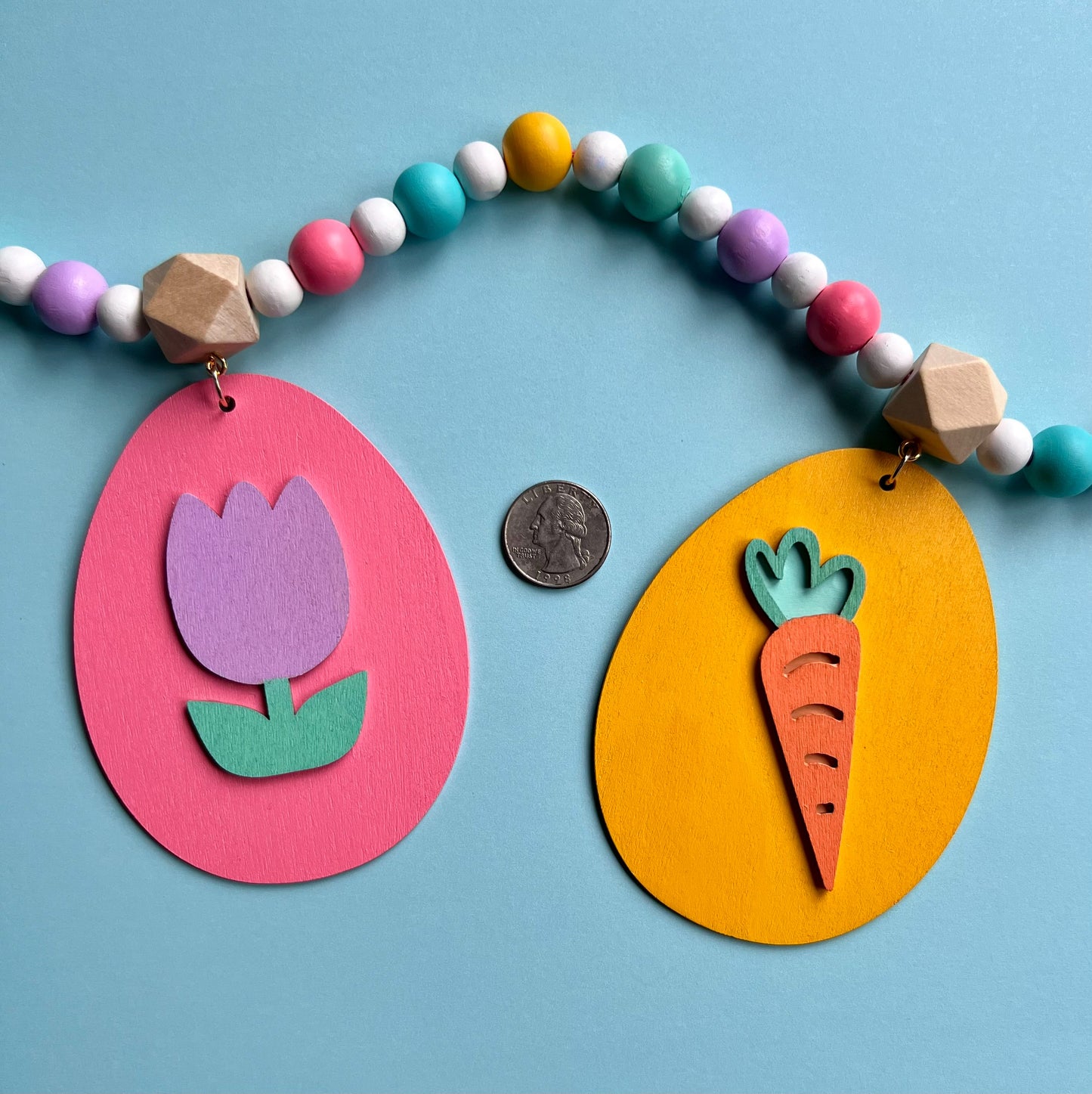 Image shows handmade garland with flat wood eggs featuring spring themed shapes and hang from a geometric wood bead. Garland has 4-inch loop on either end.