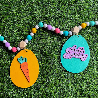 Image shows handmade garland with flat wood eggs featuring spring themed shapes and hang from a geometric wood bead. Garland has 4-inch loop on either end.