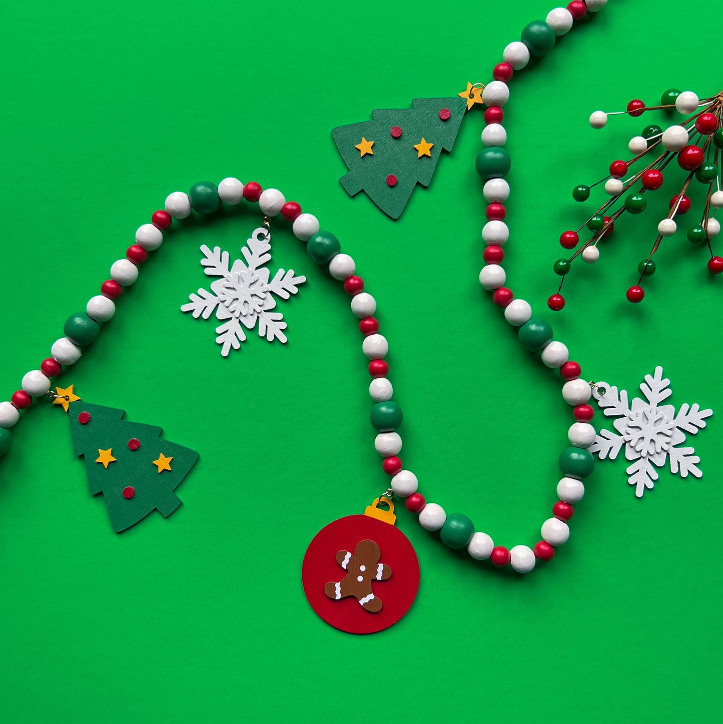 Holiday Cheer Handmade Garland. The garland features two decorated Christmas trees, two snowflakes, and one red ornament with a gingerbread man. Shapes hang from wood beads. Christmas holiday seasonal decoration or gift.