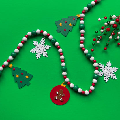 Holiday Cheer Handmade Garland. The garland features two decorated Christmas trees, two snowflakes, and one red ornament with a gingerbread man. Shapes hang from wood beads. Christmas holiday seasonal decoration or gift.