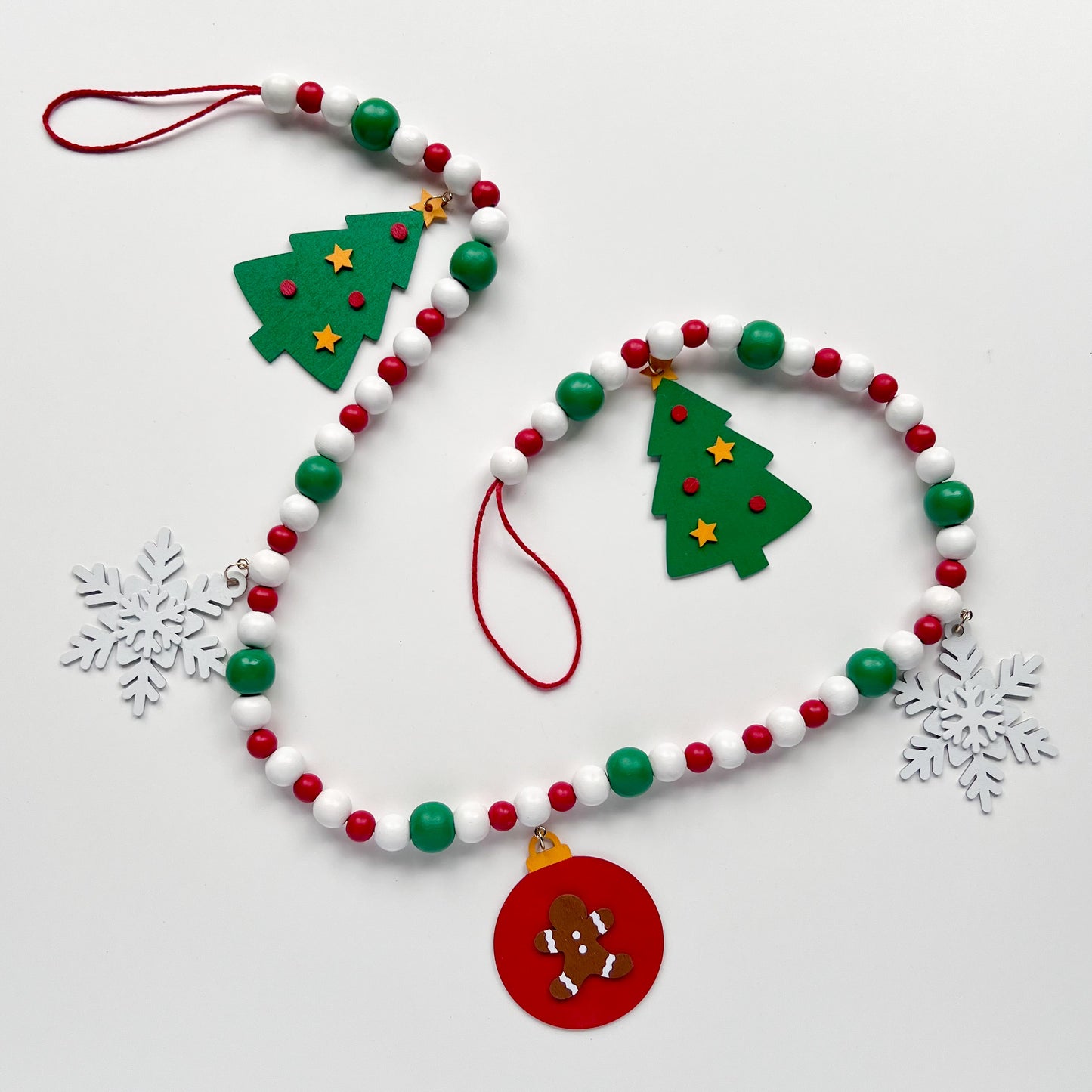 Holiday Cheer Handmade Garland. The garland features two decorated Christmas trees, two snowflakes, and one red ornament with a gingerbread man. Shapes hang from wood beads. Christmas holiday seasonal decoration or gift.