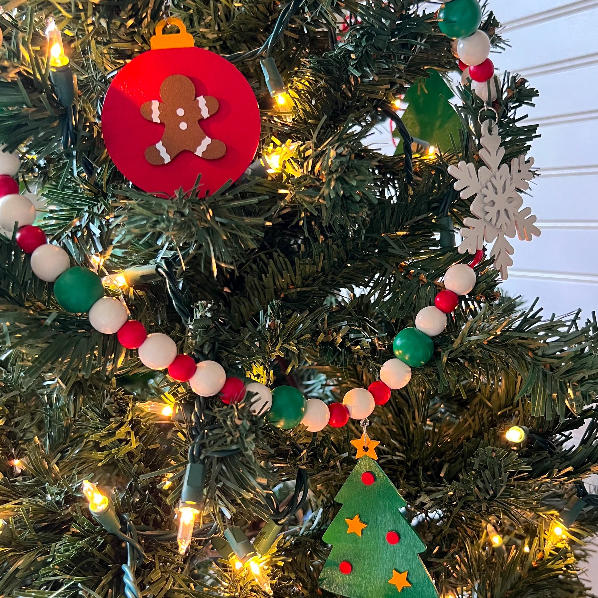 Holiday Cheer Handmade Garland. The garland features two decorated Christmas trees, two snowflakes, and one red ornament with a gingerbread man. Shapes hang from wood beads. Christmas holiday seasonal decoration or gift.