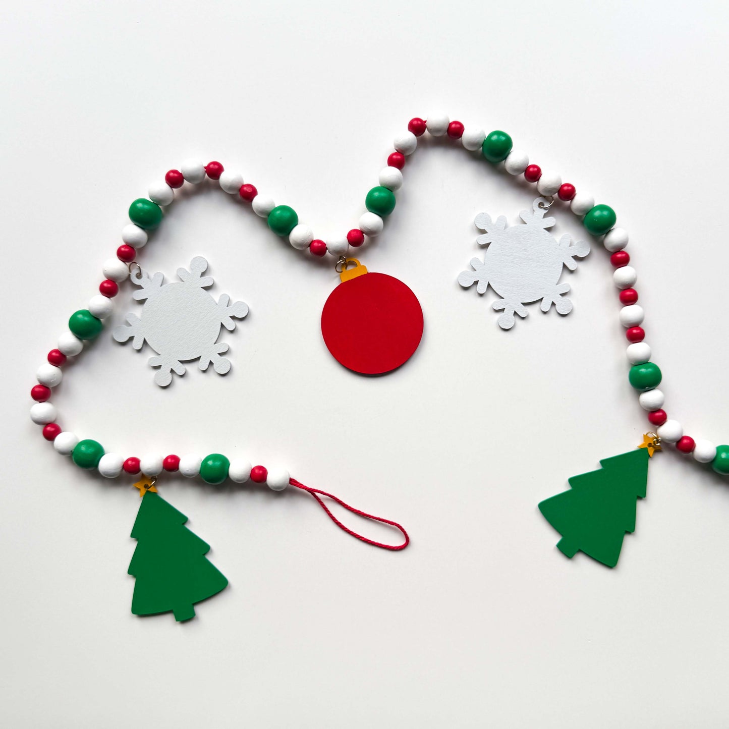 Back of a Christmas themed wood bead garland with beads painted green, red or white. 5 shapes hang from beads painted those same colors - 2 green trees, 2 white snowflakes and 1 red ornament with a gingerbread man on the front. Garland is displayed on a white background.