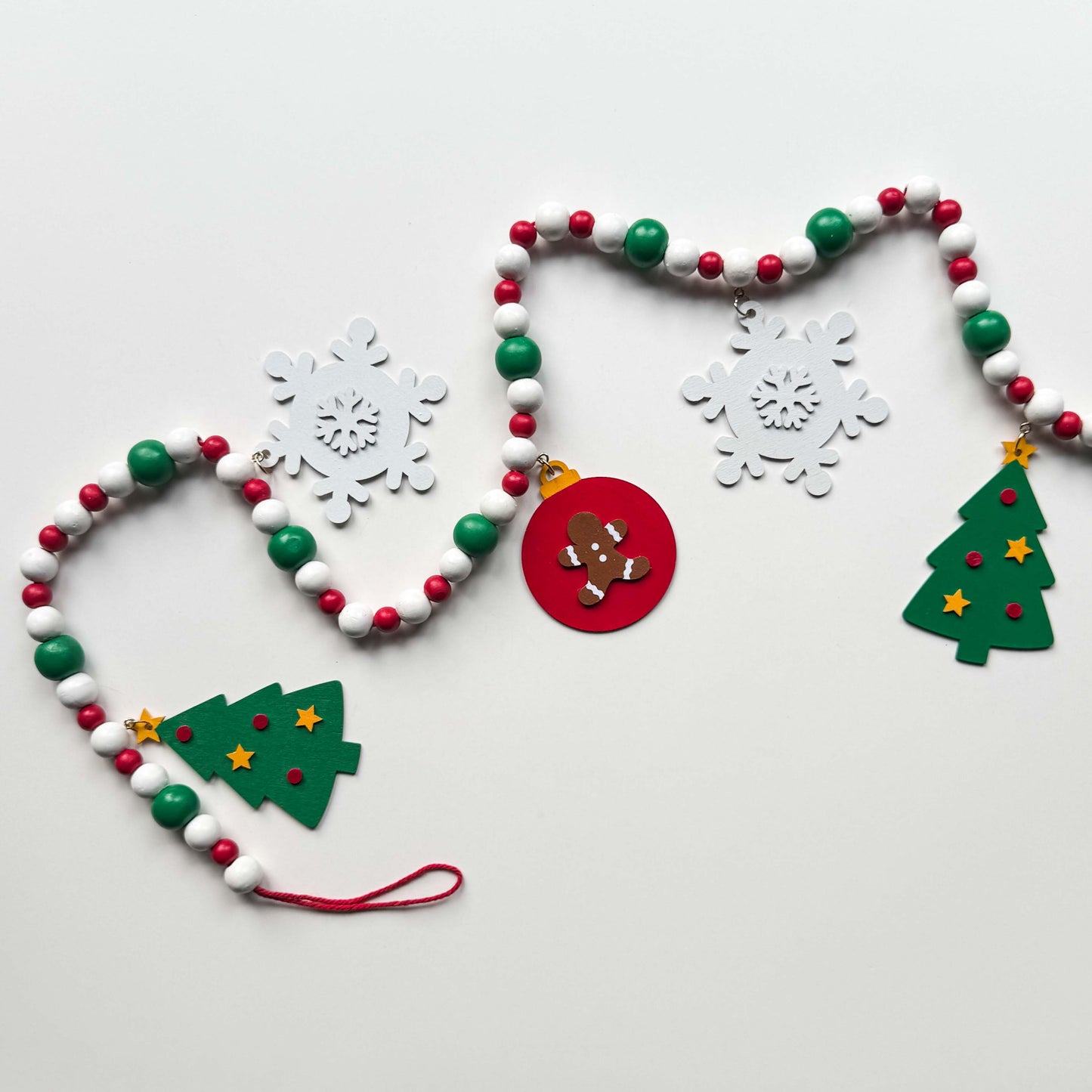 Christmas themed wood bead garland with beads painted green, red or white. 5 shapes hang from beads painted those same colors - 2 green trees, 2 white snowflakes and 1 red ornament with a gingerbread man. Garland is displayed on a white background.