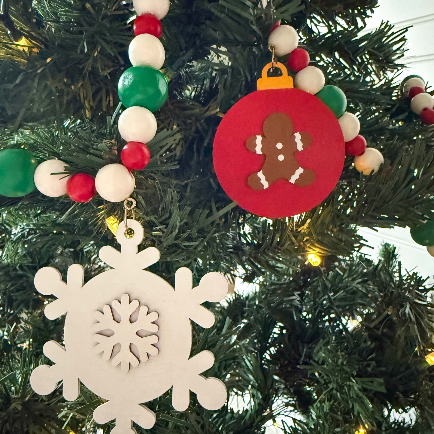 Closeup of a Christmas themed wood bead garland with beads painted green, red or white. 5 shapes hang from beads painted those same colors. The picture shows a white snowflake and a red ornament with a gingerbread man. Garland is displayed on a Christmas tree.