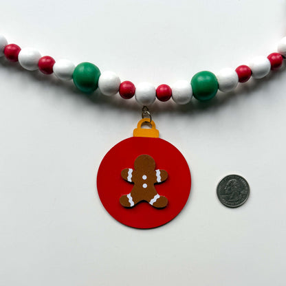 Closeup of a Christmas themed wood bead garland with beads painted green, red or white. Image shows a red ornament with a gingerbread man next to a quarter for a size comparison. Garland is displayed on a white background.