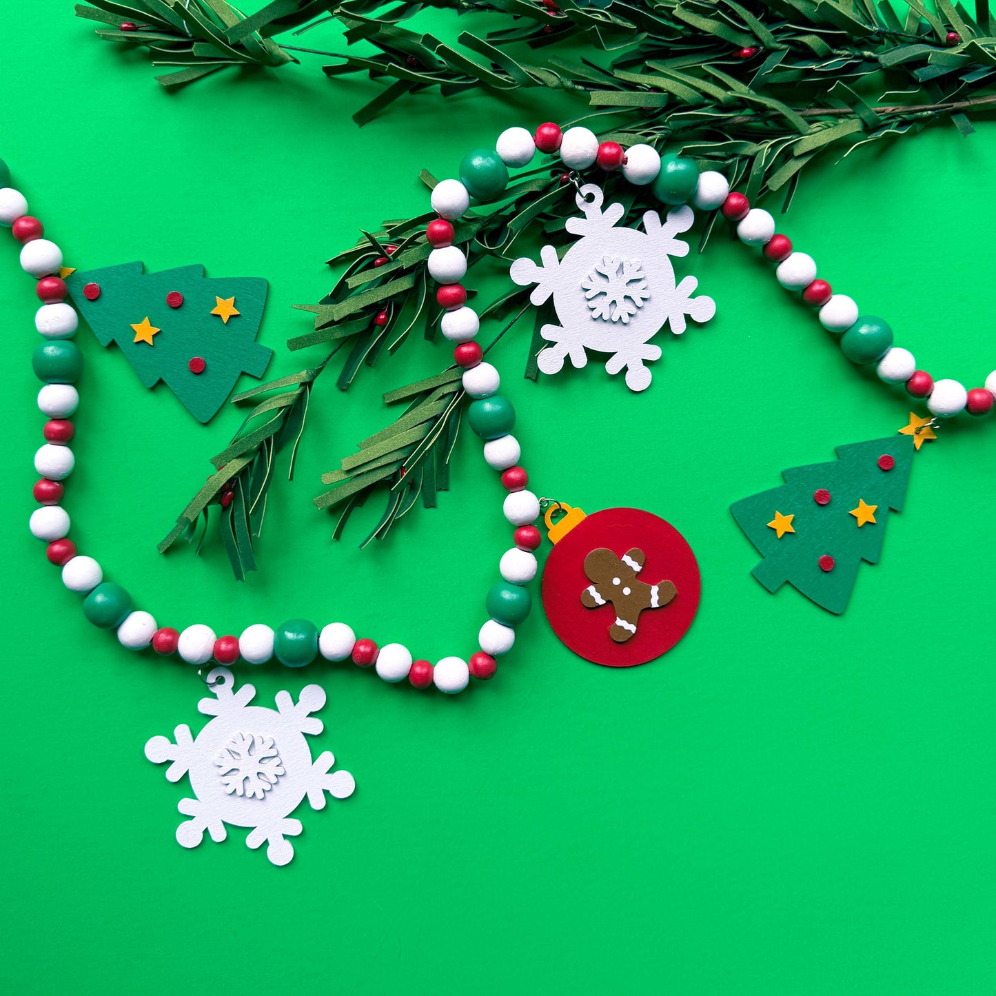 Christmas themed wood bead garland with beads painted green, red or white. 5 shapes hang from beads painted those same colors - 2 green trees, 2 white snowflakes and 1 red ornament with a gingerbread man. Garland is displayed on a green background.