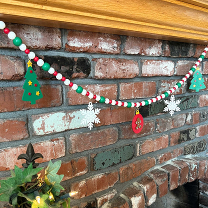 Holiday Cheer Handmade Garland. The garland features two decorated Christmas trees, two snowflakes, and one red ornament with a gingerbread man. Shapes hang from wood beads. Christmas holiday seasonal decoration or gift.