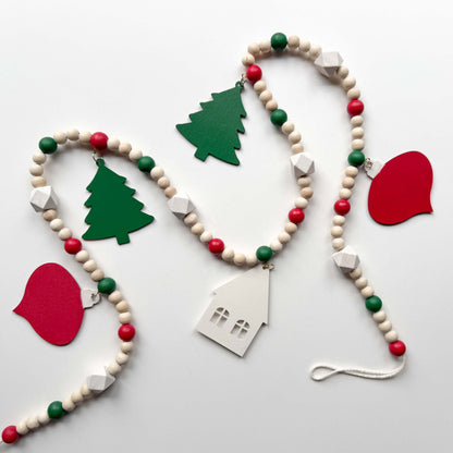 Image of the back of a Christmas wood bead garland that features two red snowflake ornaments, forest green trees, and a warm white house with a red heart. Each shape hangs from wood bead by a jump ring and eye screw. Red, warm white, green, and natural beads are strung on 2mm cream macrame cord. 
