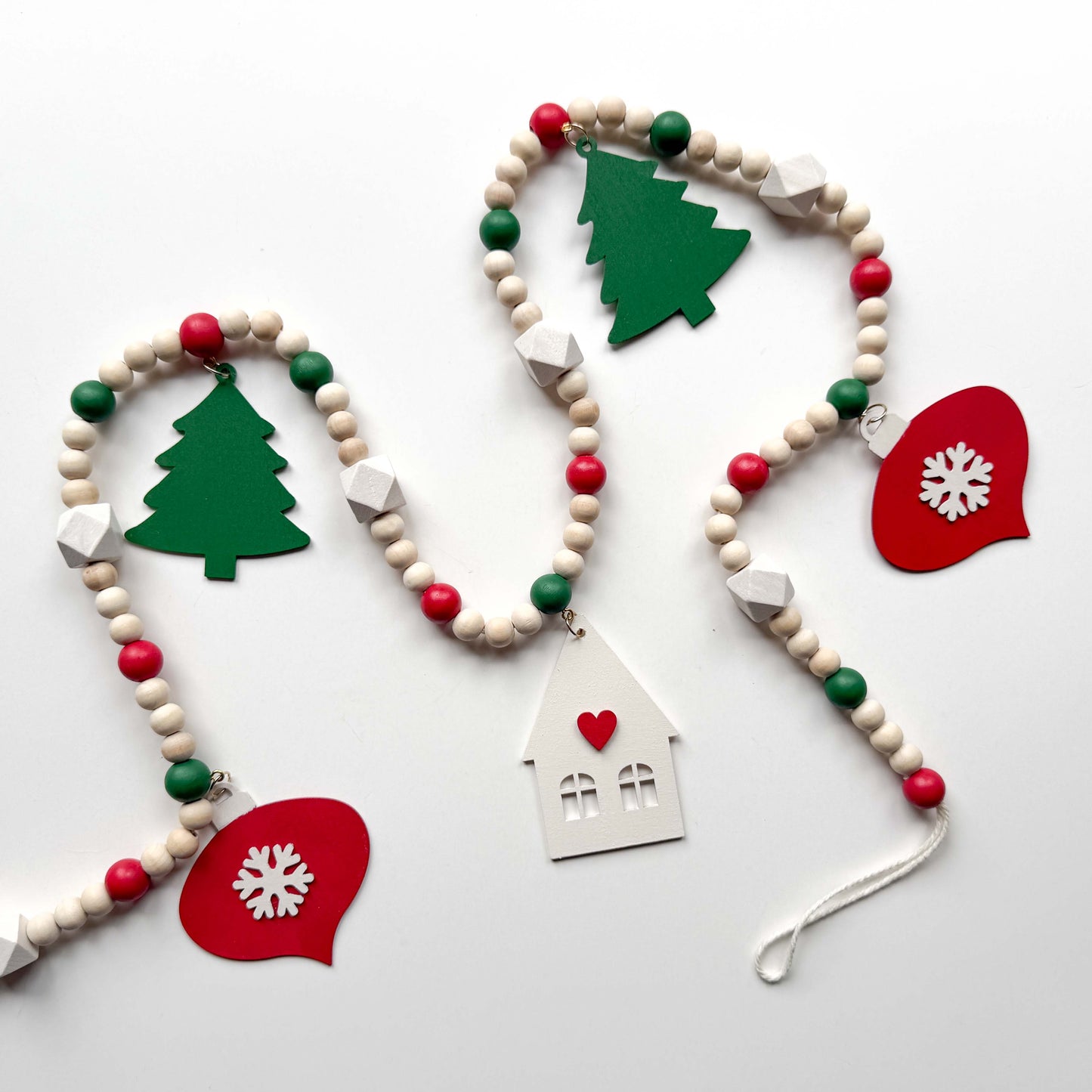 Christmas wood bead garland that features two red snowflake ornaments, forest green trees, and a warm white house with a red heart. Each shape hangs from wood bead by a jump ring and eye screw. Red, warm white, green, and natural beads are strung on 2mm cream macrame cord. 