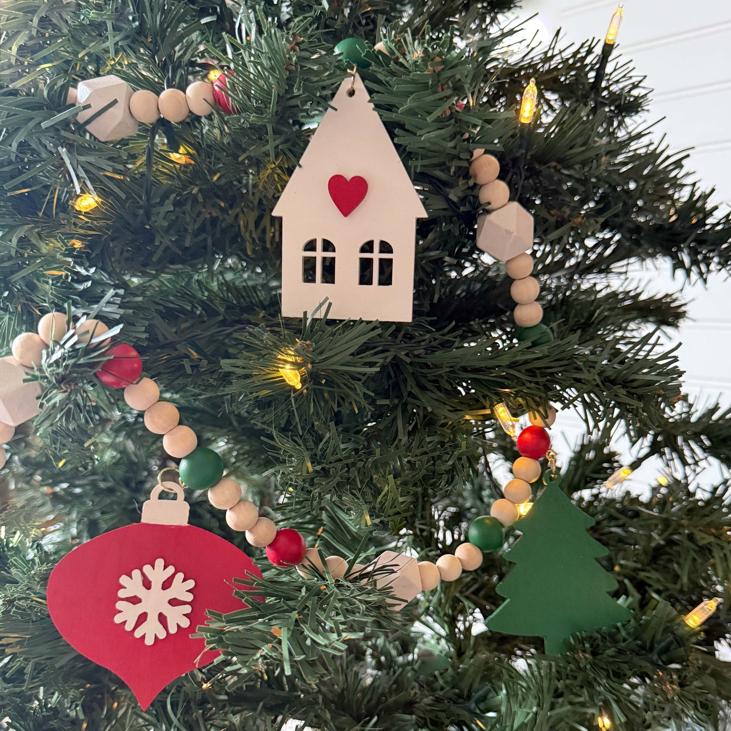 Christmas wood bead garland that features two red snowflake ornaments, forest green trees, and a warm white house with a red heart. Each shape hangs from wood bead by a jump ring and eye screw. Red, warm white, green, and natural beads are strung on 2mm cream macrame cord. Garland hangs on a Christmas tree.