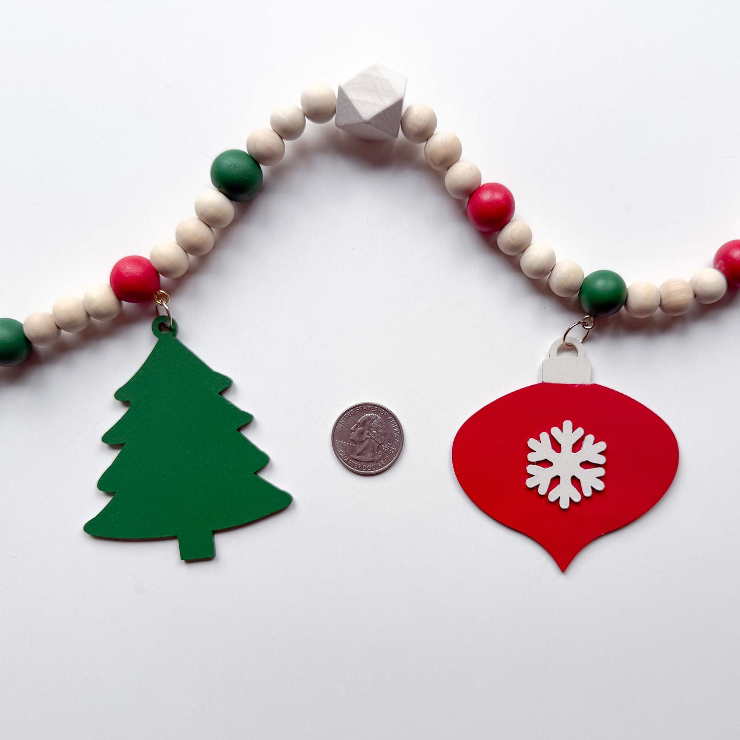 Closeup image of a Christmas wood bead garland that features a red snowflake ornament and a forest green tree. Each shape hangs from wood bead by a jump ring and eye screw. Red, warm white, green, and natural beads are strung on 2mm cream macrame cord. Garland is on a white background next to a quarter for a size comparison.
