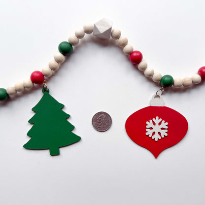 Closeup image of a Christmas wood bead garland that features a red snowflake ornament and a forest green tree. Each shape hangs from wood bead by a jump ring and eye screw. Red, warm white, green, and natural beads are strung on 2mm cream macrame cord. Garland is on a white background next to a quarter for a size comparison.