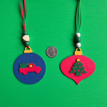 Image shows set of flat wood handmade ornaments. Each ornament features a holiday themed wood shape: a decorated Christmas tree or a truck bringing home the tree. Ornaments hang from ribbon and are decorated with three wood beads in matching colors.