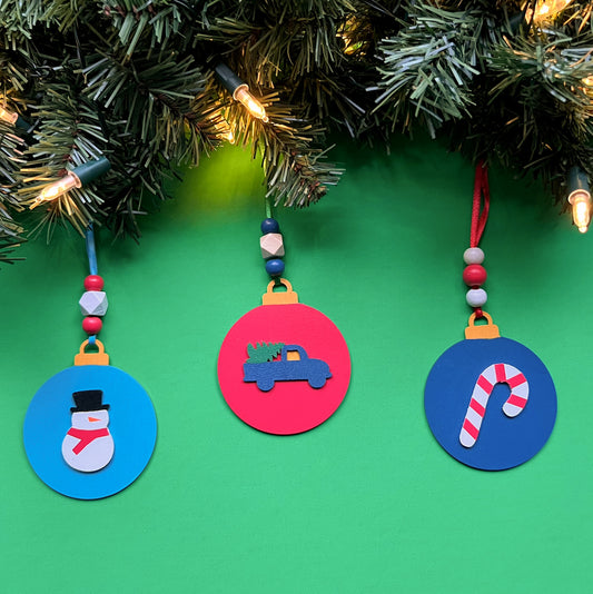 Image shows set of 3 flat wood handmade ornaments. Each ornament features a holiday themed wood shape: a snowman, a candy cane, or a truck bringing home the tree. Ornaments hang from ribbon and are decorated with three wood beads in matching colors.