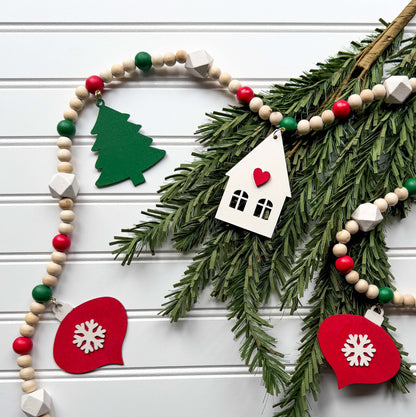 Christmas wood bead garland that features two red snowflake ornaments, forest green trees, and a warm white house with a red heart. Each shape hangs from wood bead by a jump ring and eye screw. Red, warm white, green, and natural beads are strung on 2mm cream macrame cord. 