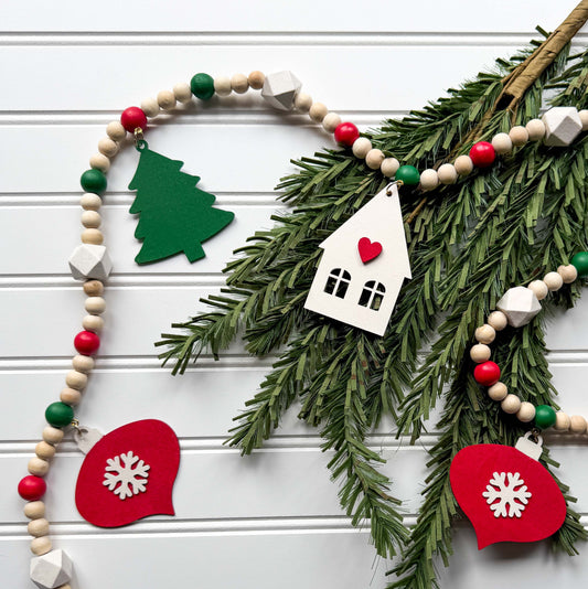 Christmas wood bead garland that features two red snowflake ornaments, forest green trees, and a warm white house with a red heart. Each shape hangs from wood bead by a jump ring and eye screw. Red, warm white, green, and natural beads are strung on 2mm cream macrame cord. 