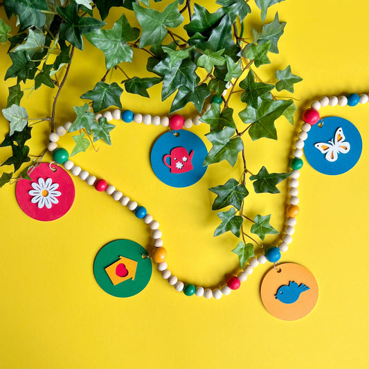 In the Garden Handmade Garland. The garland features wood circles a garden themed shape – a watering can, flower, birdhouse, bluebird, or butterfly. Shapes hang from wood beads. Spring or summer garden seasonal decoration or gift.