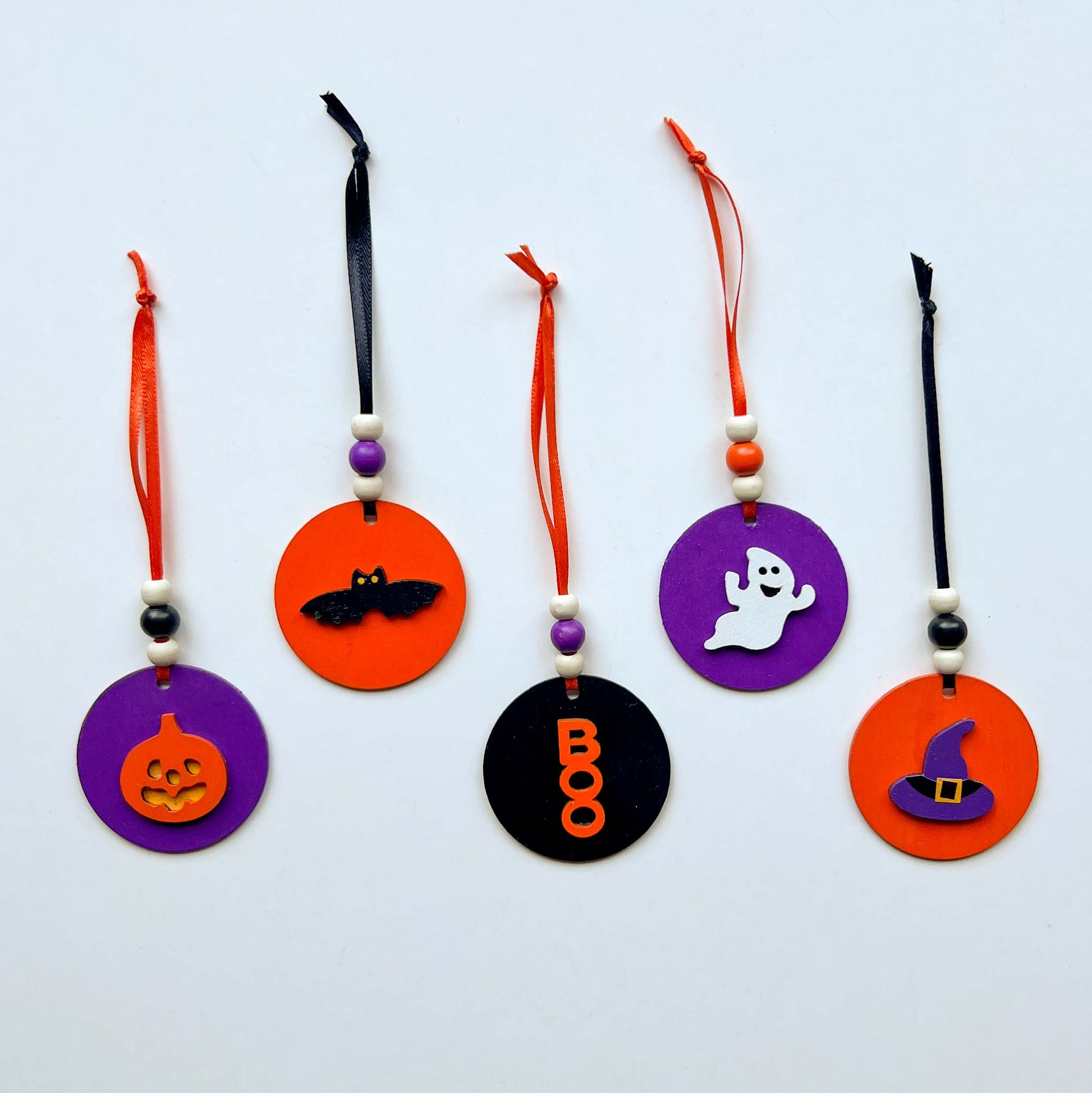 Set of 5 small wood Halloween ornaments. Feature 2 inch wood circles with Halloween themed shapes. Include three wood beads with a ribbon.